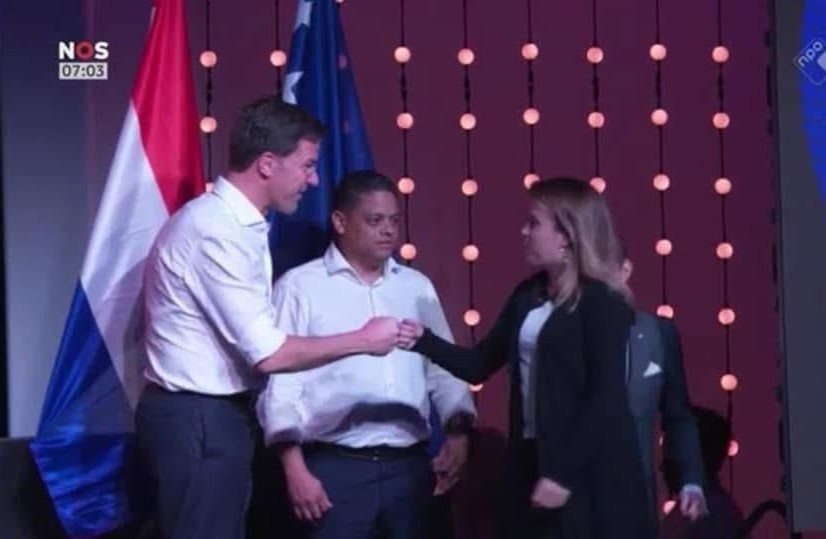 A handshake on stage in front of Dutch and Curaçao flags